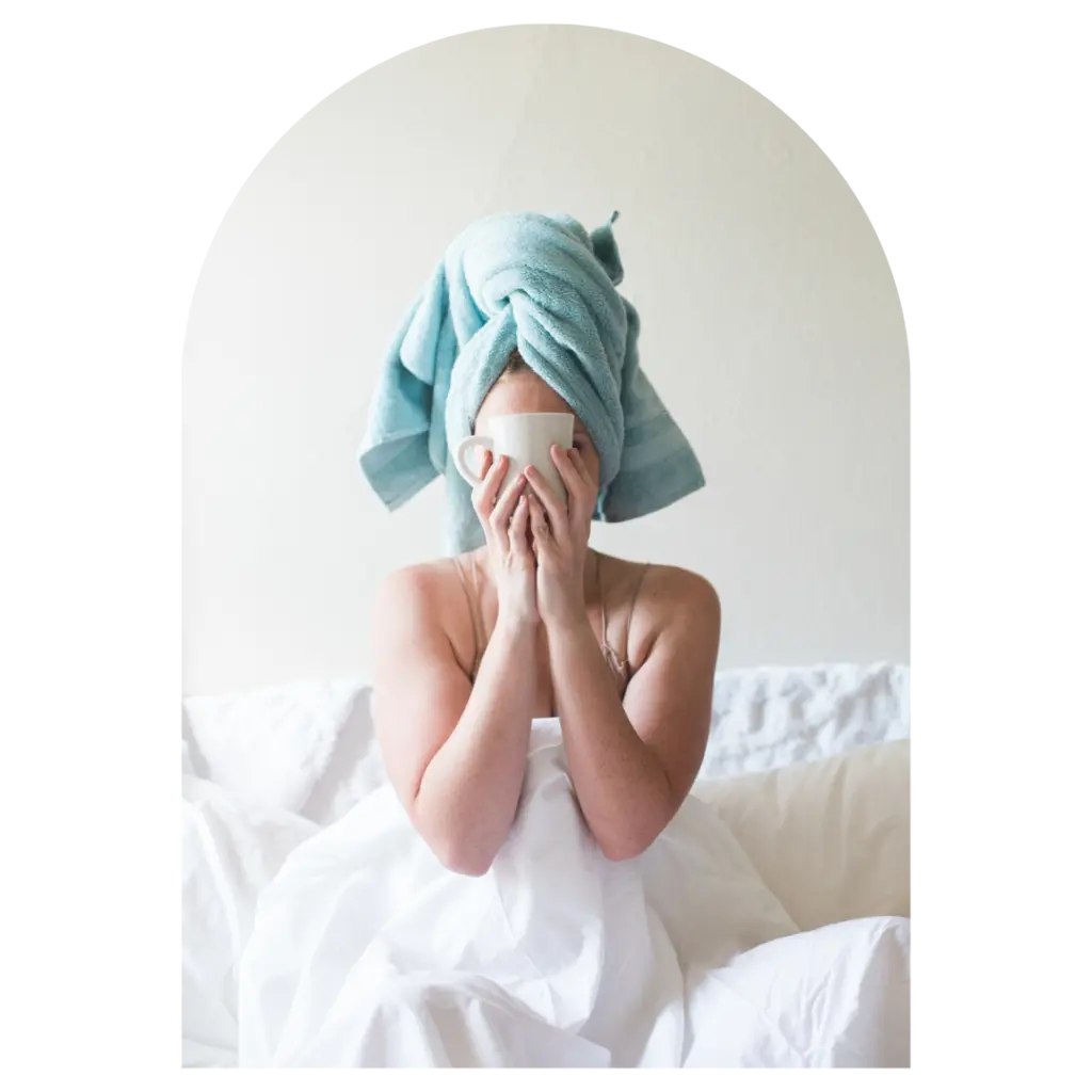 woman in bed with towel around her head holding a coffee cup in front of her face